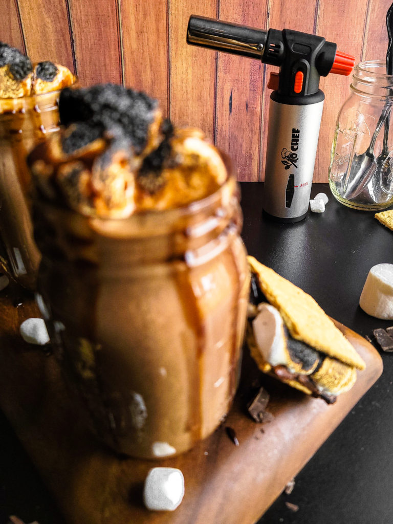 2 glasses full of S'mores hot chocolate with toasted marshmallows with a kitchen torch in the background
